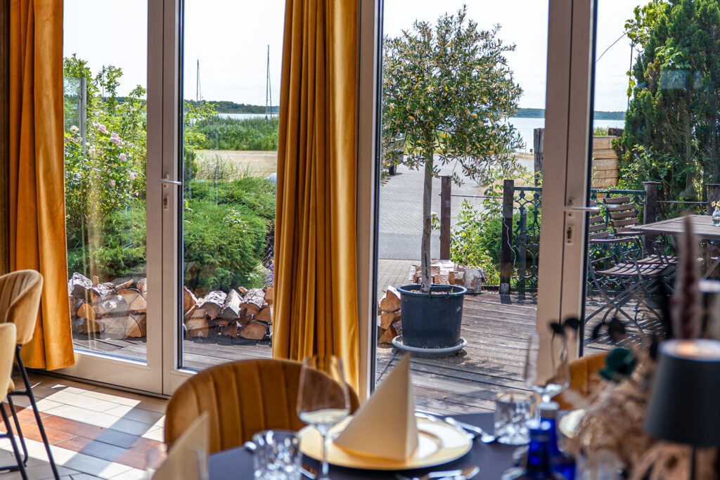 Veranstaltungssaal mit dunkler Husse, goldenen Platztellern, Tischschmuck. Im Hintergrund bodentiefe Fenster mit Blick auf den Außenbereich und See