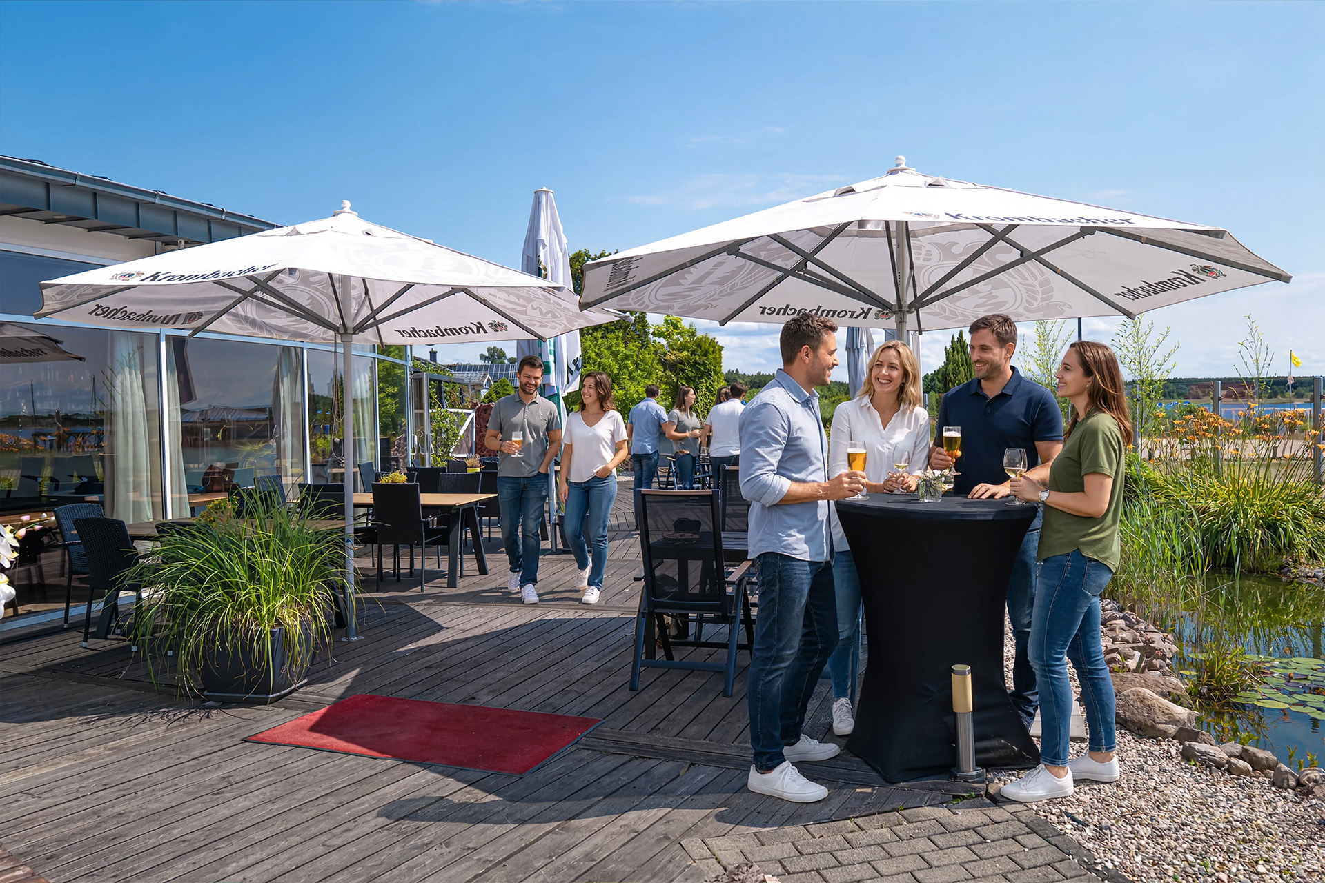 Terrasse der Eventlocation mit Holzdeck, Sonnenschirmen, Sitzgruppen. Mehrere Personen stehen hier zusammen und trinken etwas