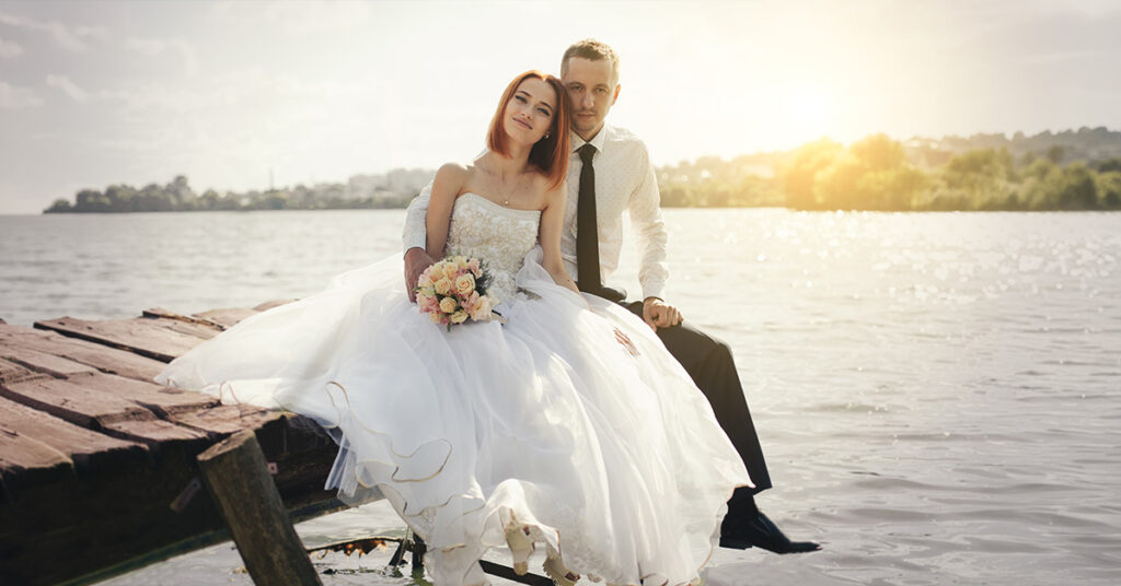 Ein Hochzeitspaar sitzt am Tag der Hochzeit bei Sonnenuntergang auf einer Brücke in der Nähe eines Sees.